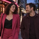A smiling adult couple walking past a cinema at dusk in pink and plum clothing